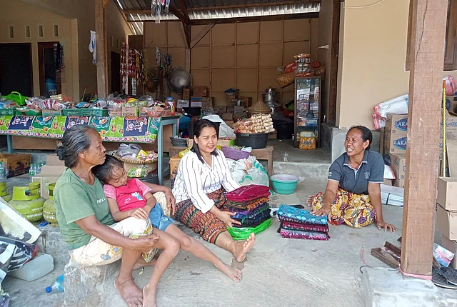 This image shows several people, including women and a child, gathered and sitting together in what appears to be a small local shop or porch area in a rural Southeast Asian setting.