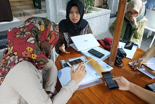 This image shows young women sitting at a wooden table, working on documents and interacting via digital devices such as smartphones and a laptop.