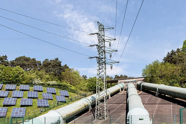 The image shows a pumped-storage power station with its distinctive large pressure pipelines, a high-voltage power line and an adjacent photovoltaic plant.